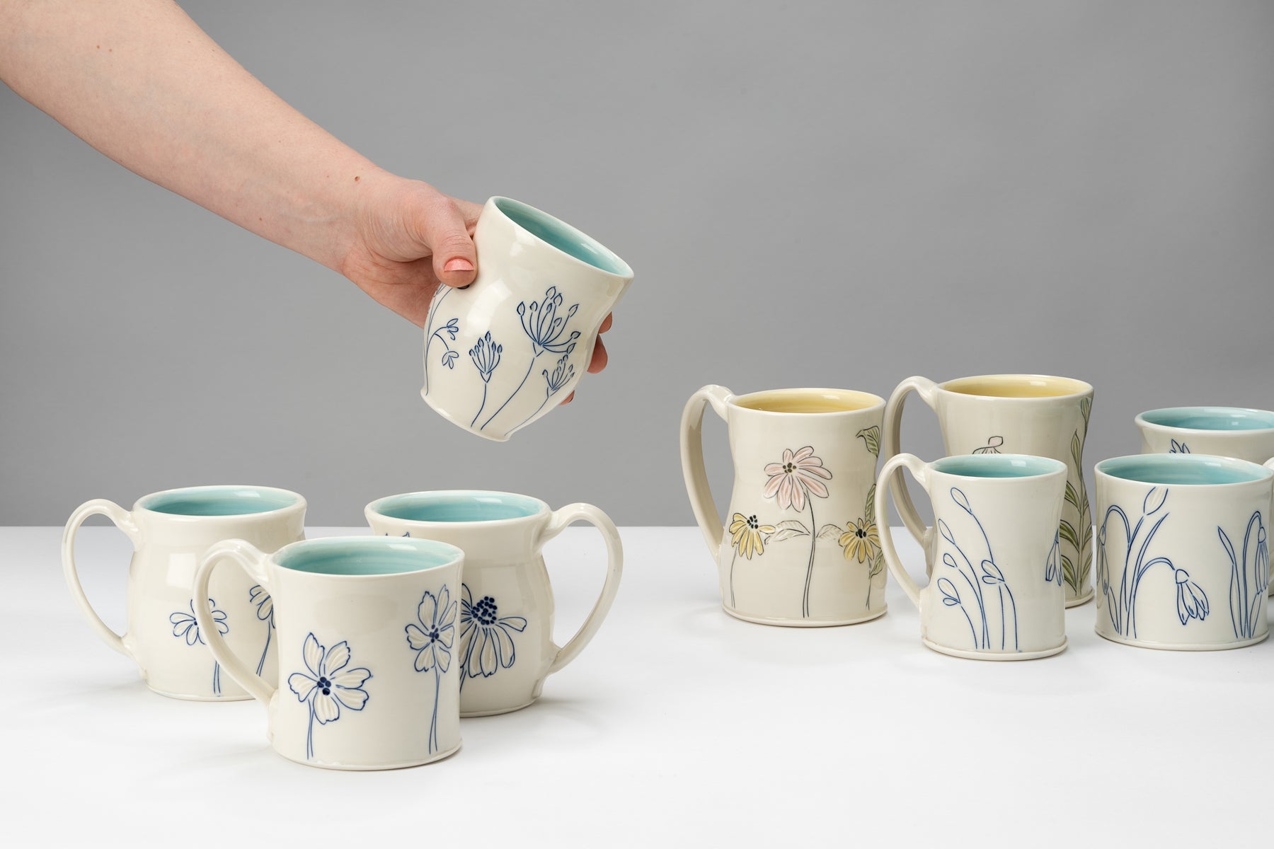 White and blue handmade porcelain cups and mugs by ceramic artist Julie Wiggins sit on a white table against a grey background. Image by Loam