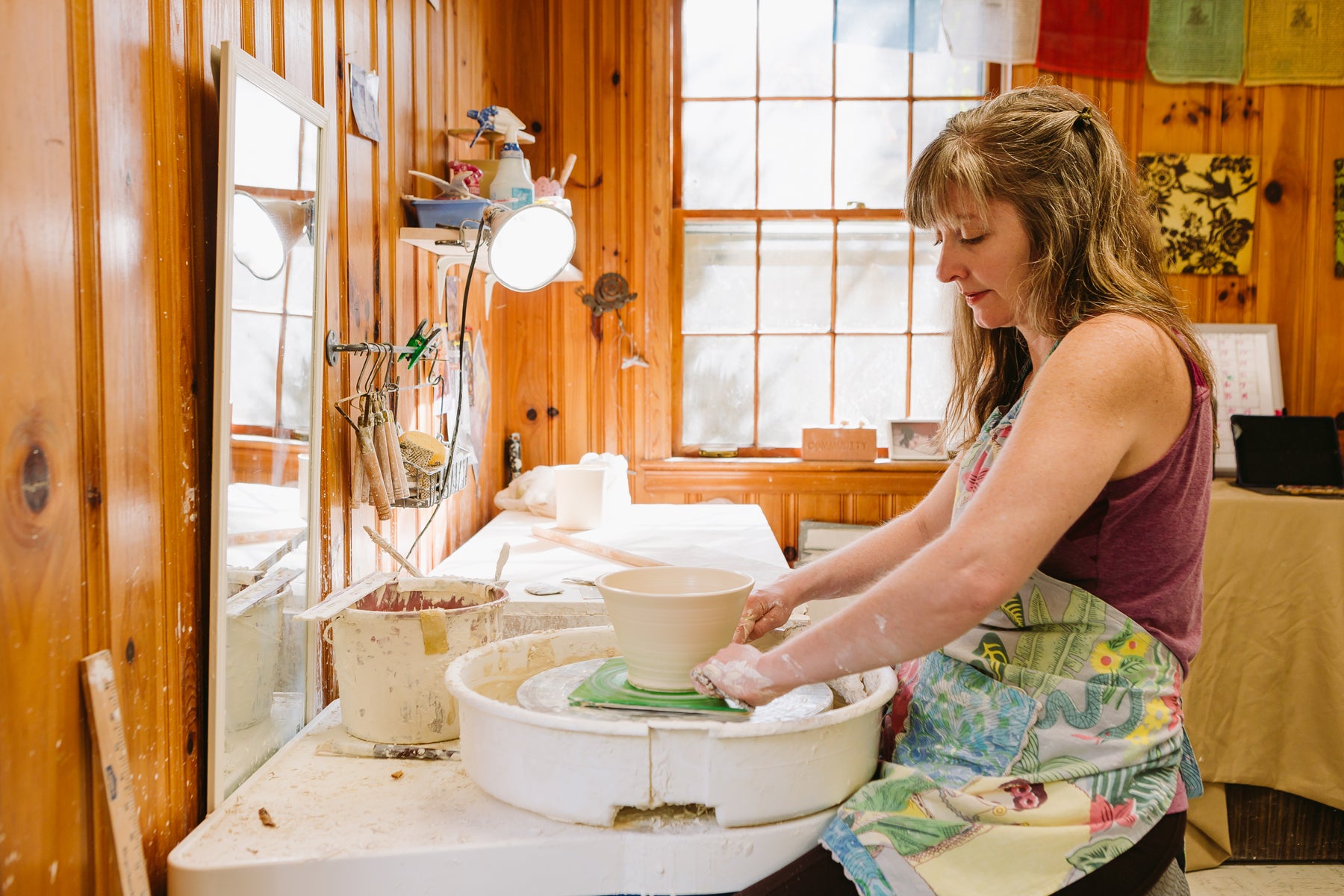 Ceramic artist Julie Wiggins sitting at her potters' wheel in her wood paneled studio, the side of her face towards the camera