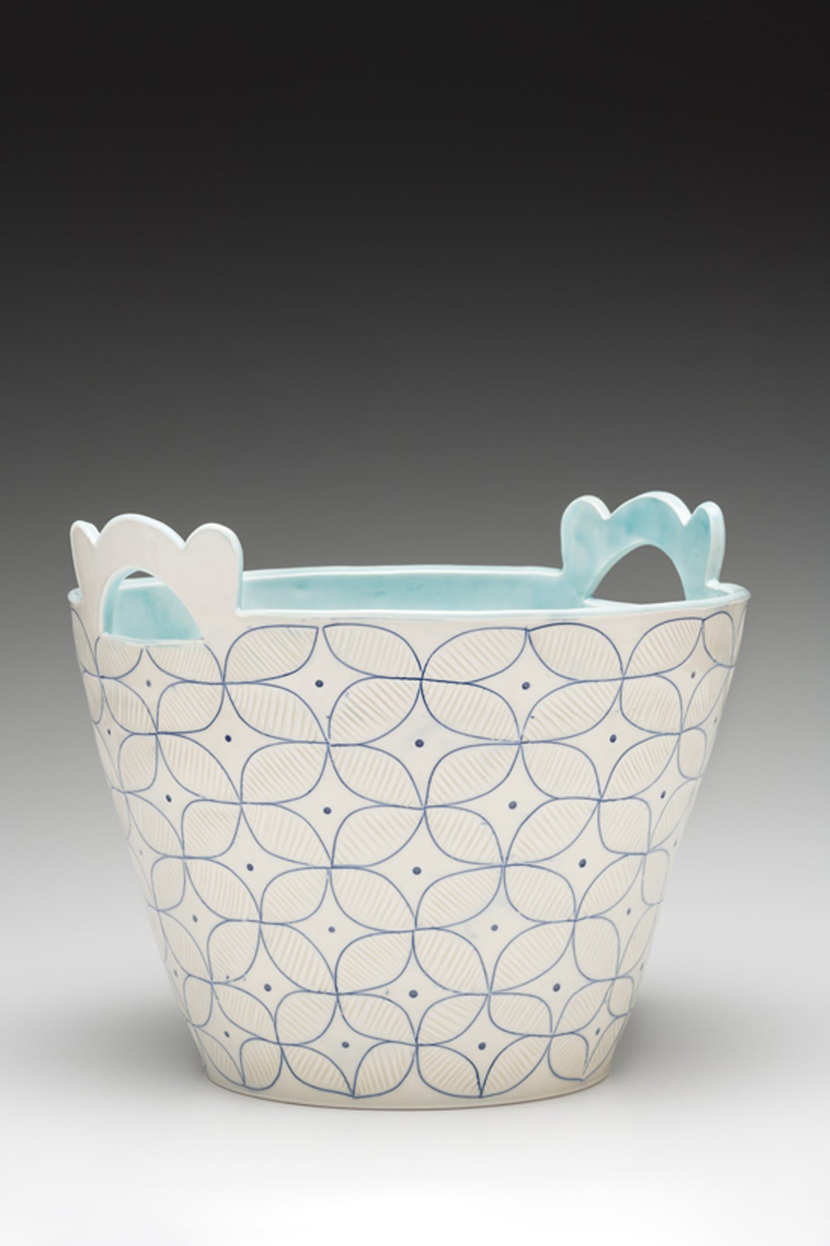 A white handmade porcelain basket vessel with blue inlay by ceramic artist Julie Wiggins sits against a gradient background. 