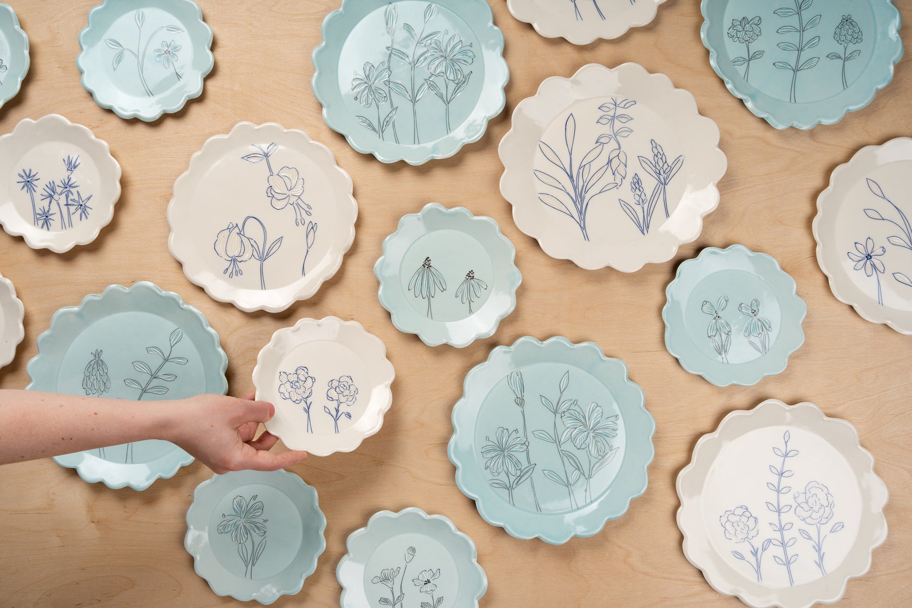 A field of white and blue floral scalloped porcelain plates by potter Julie Wiggins. A hand reaches into the frame to set down a small plate. Image by Loam.