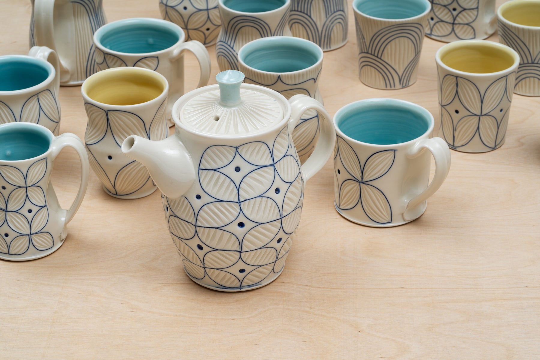 A white porcelain teapot with blue geometric designs made by ceramic artist Julie Wiggins surrounded by matching cups with blue and yellow glazed interiors. Image by Loam