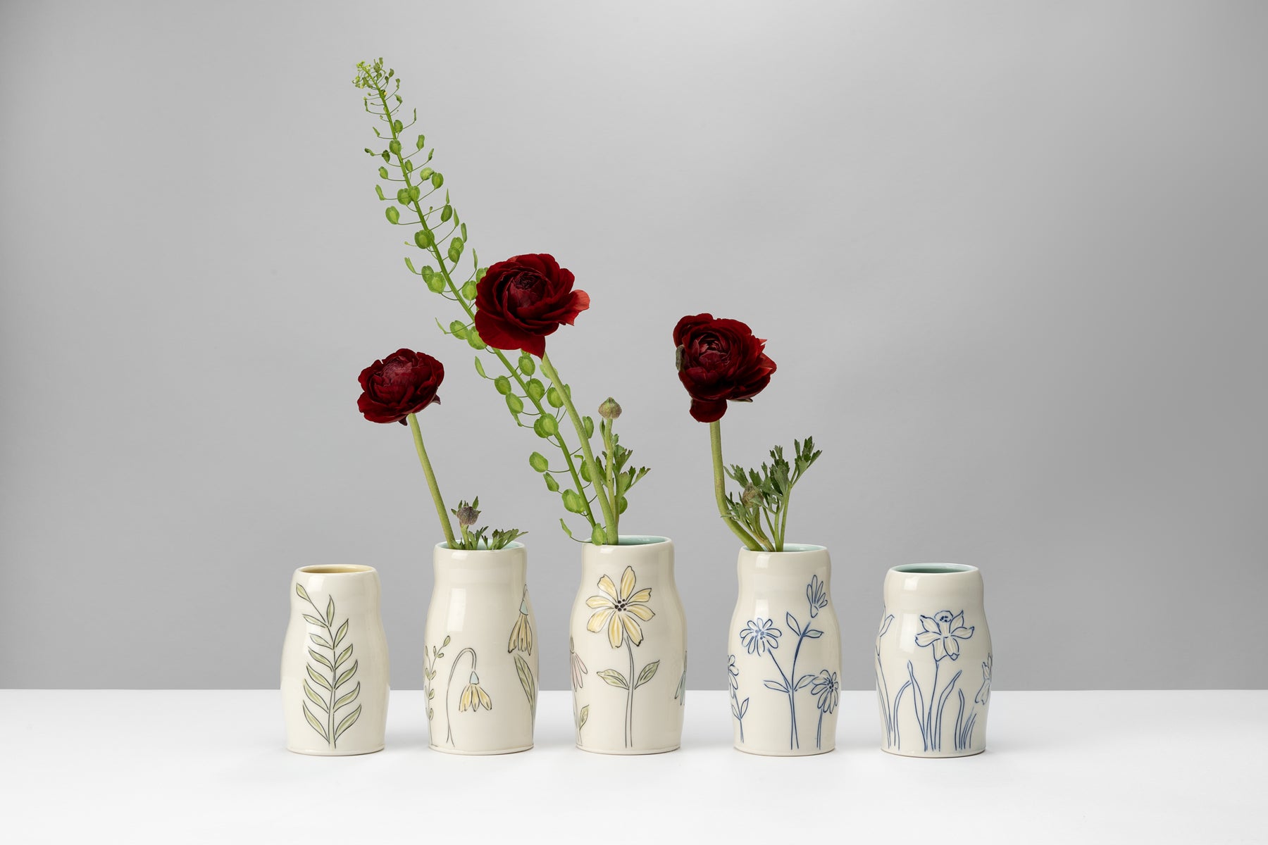 White and blue handmade porcelain pottery pieces by ceramic artist Julie Wiggins sit on a white table against a grey background and hold deep red flowers. Image by Loam