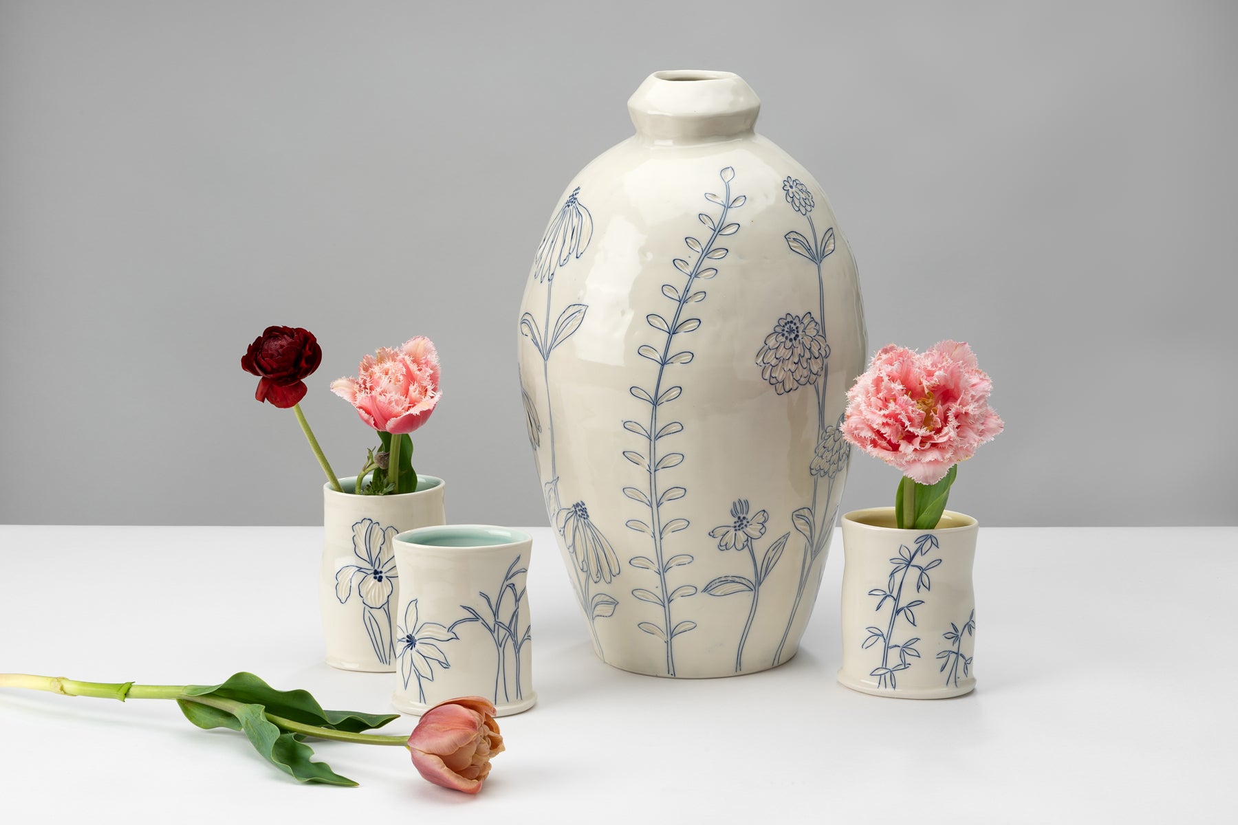White and blue handmade porcelain pottery pieces by ceramic artist Julie Wiggins sit on a white table against a grey background and hold deep red and light pink flowers. Image by Loam