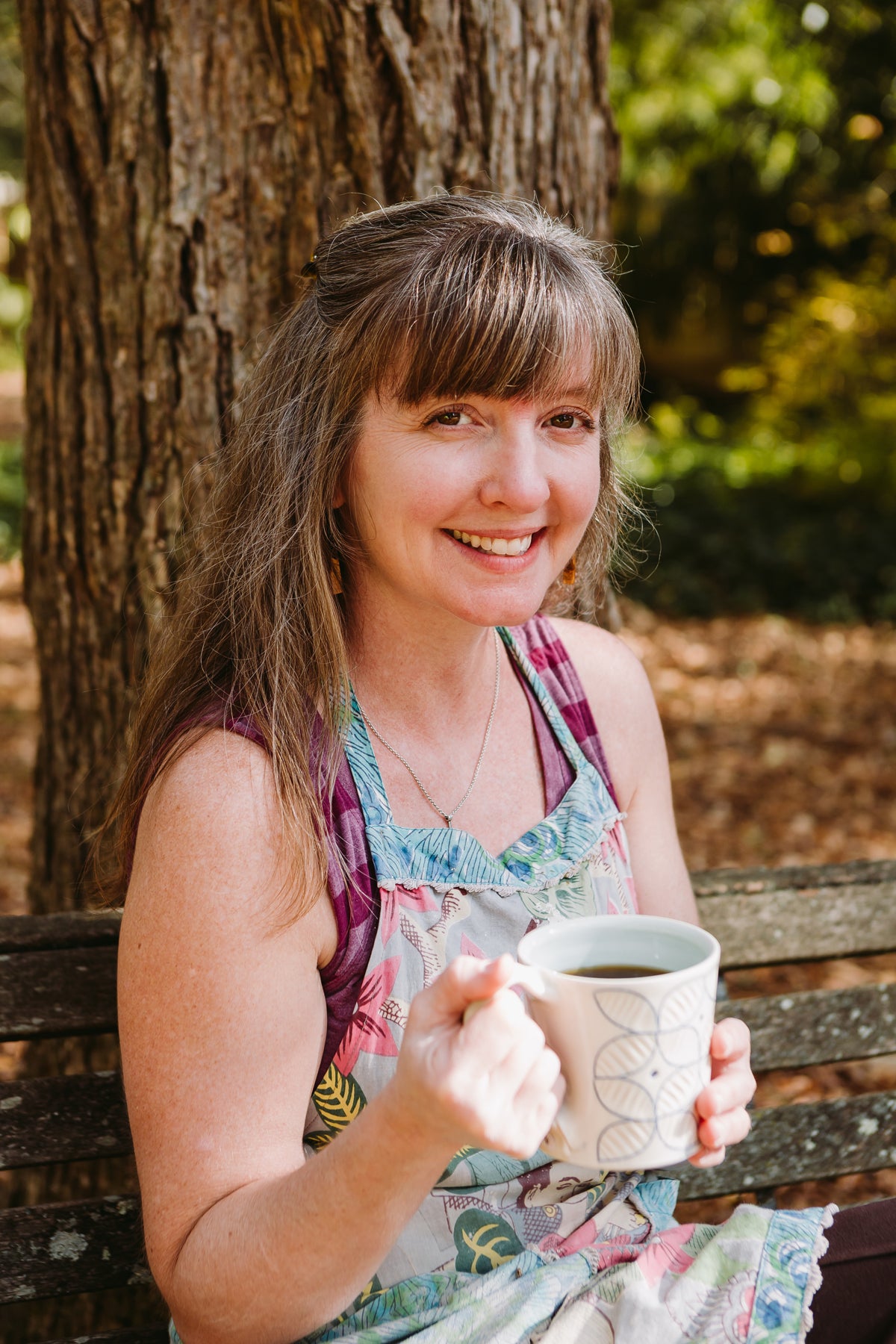 Julie Wiggins from the waist up sitting on a park bench outside holding a porcelain handmade mug that she made 