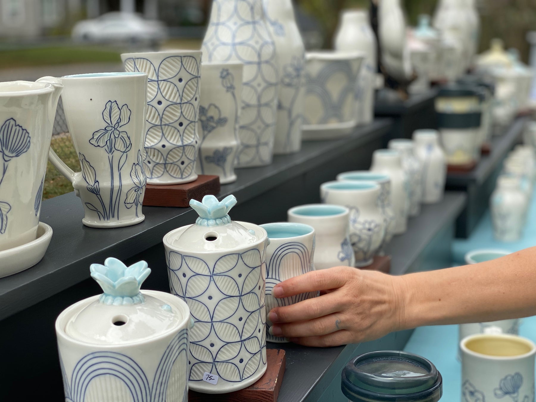 Handmade porcelain pottery pieces by artist Julie Wiggins are lined up on shelves and blue into the distance as a hand reaches to grab a cup in the foreground. 