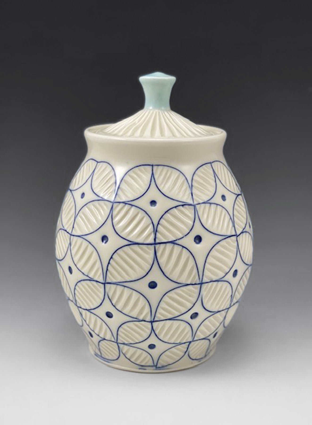 A white handmade porcelain jar with blue inlay by ceramic artist Julie Wiggins sits against a gradient background. 
