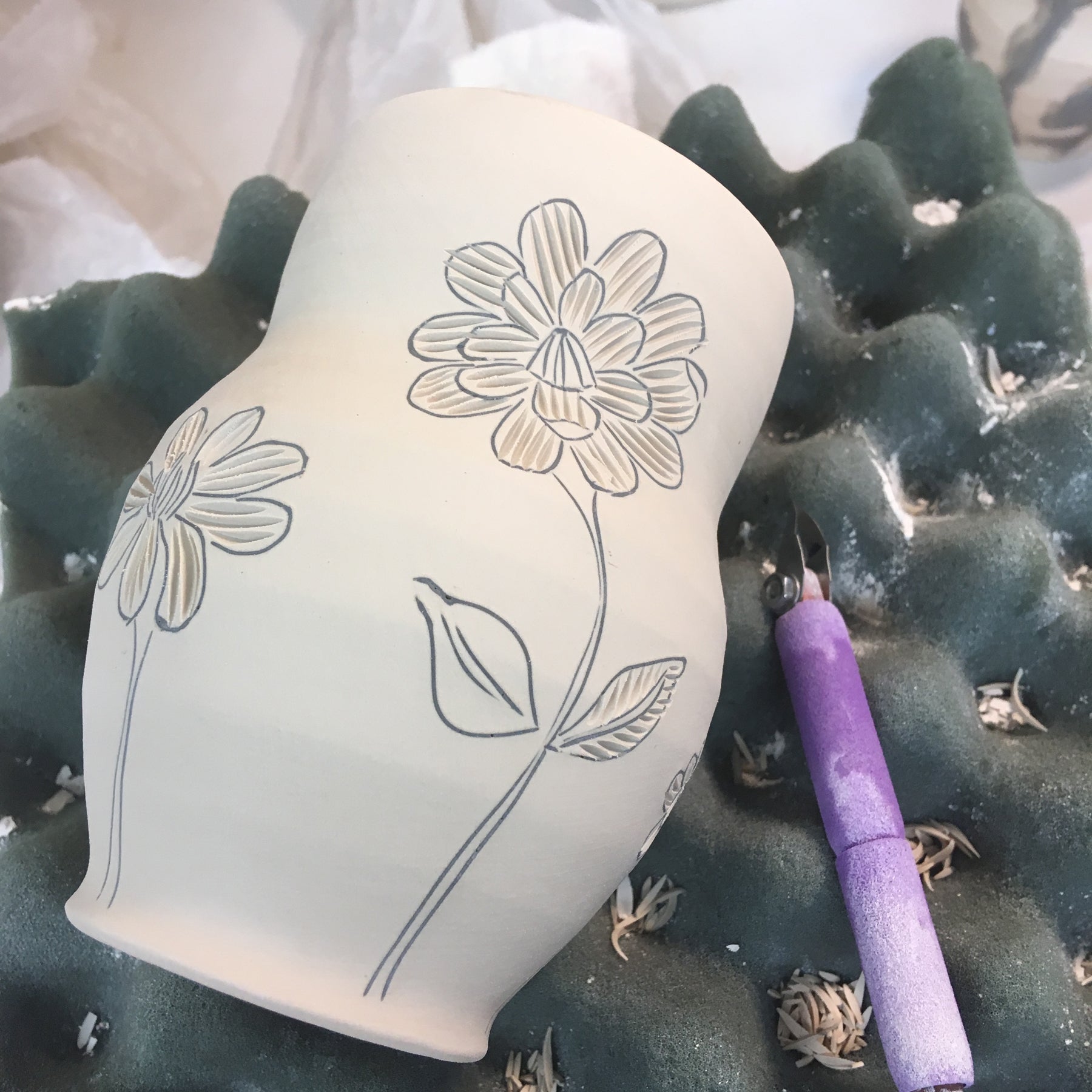 An in progress porcelain cup in sitting on foam with a carving tool laying next to it. the cup has a floral pattern carved into it.