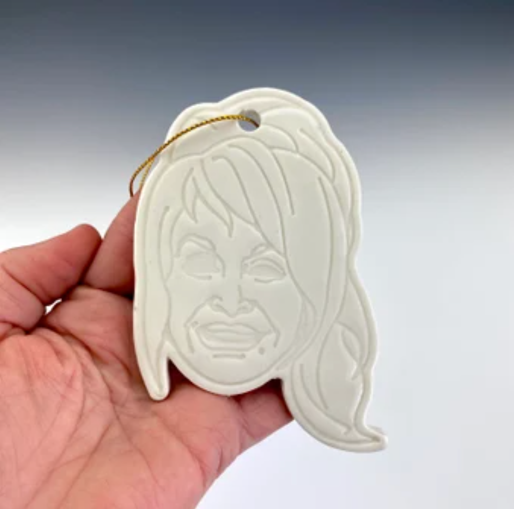 A white ornament in the shape of Dolly Parton's silhouette with a ponytail by ceramic artist Julie Wiggins against a gradient background