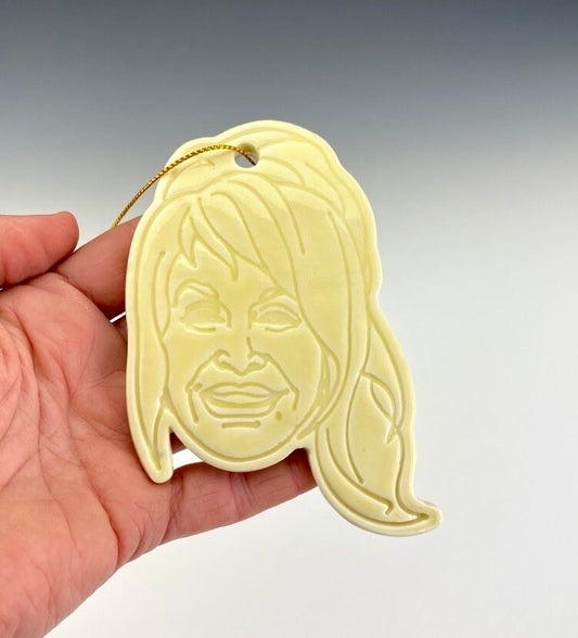 A yellow ornament in the shape of Dolly Parton's silhouette with a ponytail by ceramic artist Julie Wiggins against a gradient background